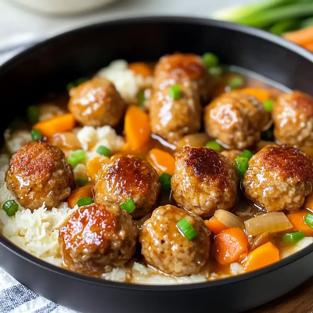 A dark bowl showing tender meatballs in a savory, creamy sauce with carrots and onions, all over cauliflower rice garnished with green onions.