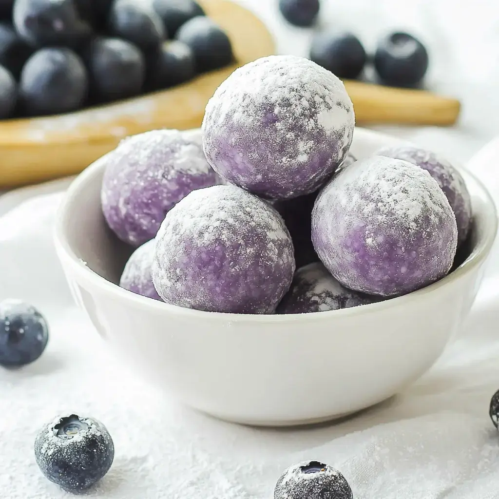 A bowl of round purple treats dusted with powdered sugar, sitting on a white table near loose blueberries.